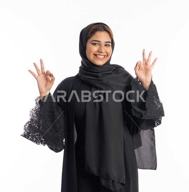 مفهوم الموافقة و القبول، علامة الامتياز و الجودة، بورتريه مقرب لامراة عربية خليجية اماراتية مبتسمة محجبة ترتدي العباءة السوداء  ترفع يدها بإيماءات تدل على الإعجاب والرضى، خلفية بيضاء