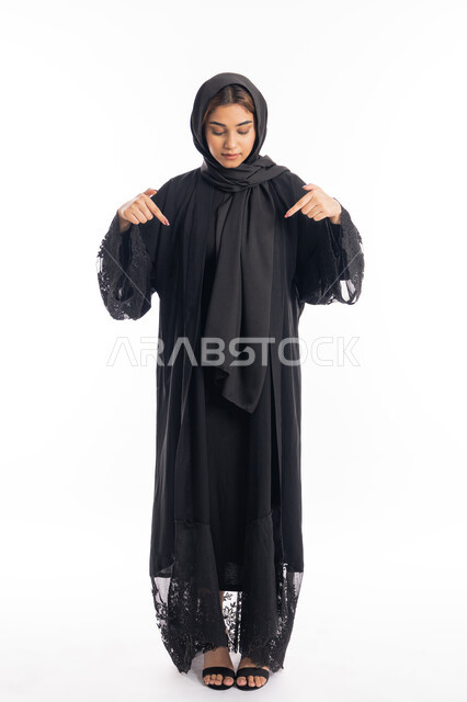 الوقوف باستقامة والاشارة الى شيء ما، تحديد موقع شيء معين، بورتريه مقرب لامراة عربية خليجية اماراتية مبتسمة محجبة ترتدي العباءة السوداء تشير بإصبعي السبابة إلى جهة الأسفل، الجسم بالطول الكامل، خلفية بيضاء