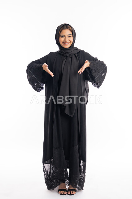 حركات تدل على المكان، التأكيد على أمر محدد, فتح اليدين لتحديد شيء ما, الاناقة و الاهتمام بالمظهر الخارجي، بورتريه لامراة عربية خليجية اماراتية مبتسمة محجبة ترتدي العباءة السوداء تشير بيديها الى مكان ما، الجسم بالطول الكامل، خلفية بيضاء