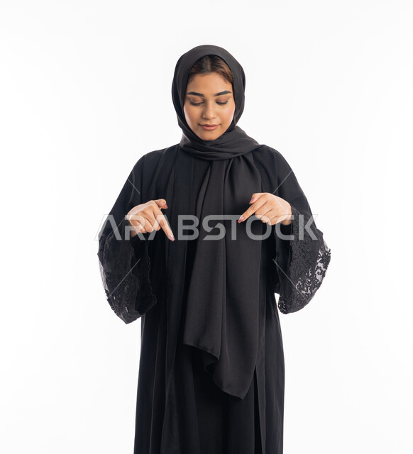 الاشارة الى شيء ما بهذا المكان، تحديد موقع شيء معين، بورتريه مقرب لامراة عربية خليجية اماراتية مبتسمة محجبة ترتدي العباءة السوداء تشير بإصبعي السبابة إلى جهة الأسفل، خلفية بيضاء