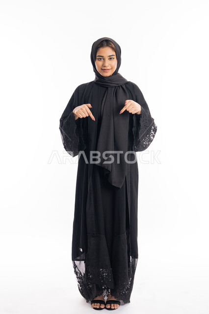 تحديد موقع شيء معين، إشارات وحركات يدين تدل على هذا المكان (هنا)، بورتريه لامراة عربية خليجية اماراتية مبتسمة محجبة ترتدي العباءة السوداء تشير بإصبعي السبابة إلى جهة الأسفل، الجسم بالطول الكامل، خلفية بيضاء