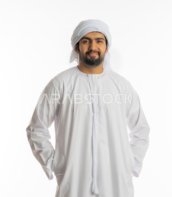 الوقوف باستقامة والنظر الى الكاميرا بتعابير السرور والرضى، مفهوم الاناقة والاهتمام بالمظهر الخارجي، بورتريه لرجل عربي خليجي إماراتي يرتدي الكندورة و الغترة البيضاء يضع يديه في جيبه، خلفية بيضاء