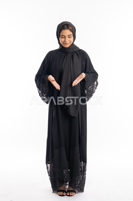 فتح اليدين لتحديد شيء ما, الاناقة و الاهتمام بالمظهر الخارجي، بورتريه لامراة عربية خليجية اماراتية مبتسمة محجبة ترتدي العباءة السوداء تشير بيديها الى مكان ما، الجسم بالطول الكامل، خلفية بيضاء