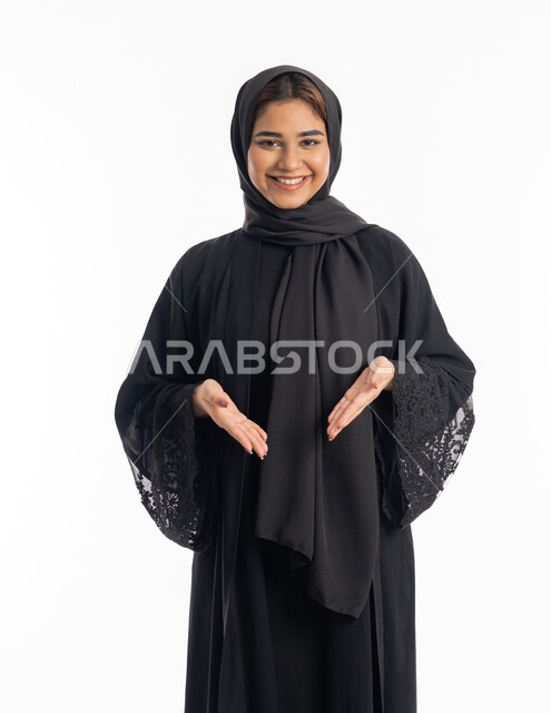 Emphasizing a specific matter, opening the hands to identify something, elegance and attention to external appearance, a close-up portrait of a smiling, veiled Arab Gulf Emirati woman wearing a black abaya, pointing with her hands to a place, white background.