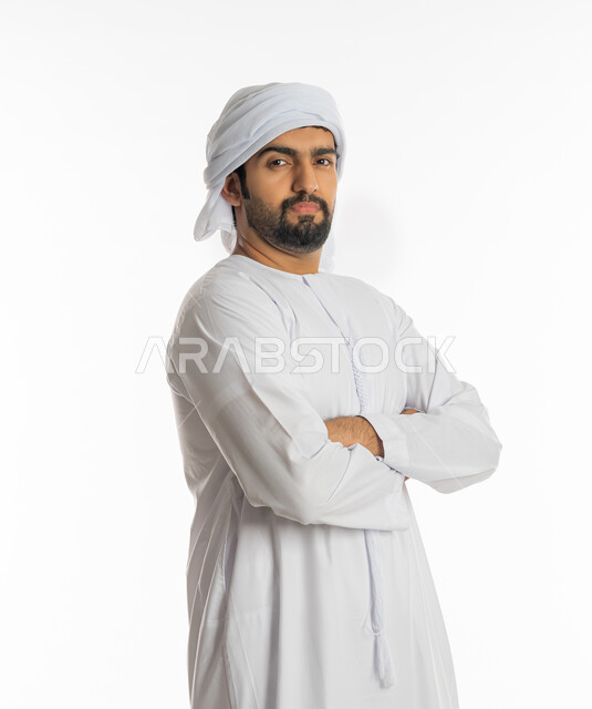 الوقوف بإستقامة متكتف اليدين، إيماءات الشعور بعدم الرضا والانزعاج, بورتريه لرجل عربي خليجي إماراتي يرتدي الكندورة و الغترة البيضاء يقف بثقة وينظر الى الكاميرا،  خلفية بيضاء