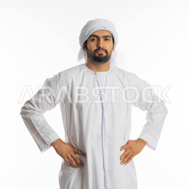 Gestures of dissatisfaction and annoyance, standing straight and placing hands on the waist, portrait of an Emirati Gulf Arab man wearing a kandora and white ghutra, standing confidently and looking at the camera, white background