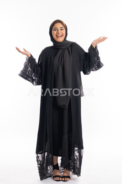 الوقوف بثقة و استقامة، الاناقة و الاهتمام بالمظهر الخارجي، بورتريه لامراة عربية خليجية اماراتية مبتسمة محجبة ترتدي العباءة السوداء تفتح يديها بايماءات الرضا و السرور، الجسم بالطول الكامل، خلفية بيضاء