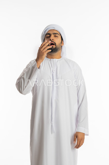 الإحساس بالإرهاق و الكسل، الشعور بالتعب والتثاؤب، الحاجه الى النوم والراحه, بورتريه مقرب لرجل عربي اماراتي خليجي يرتدي الكندورة والعمامة يضع يده على فمه ،إيماءات النعاس والخمول , خلفية بيضاء