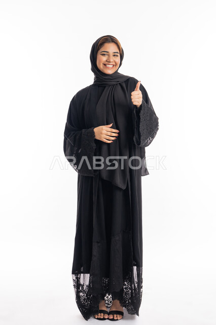 Elegance and attention to external appearance, feeling good, the concept of approval and acceptance, portrait of a smiling Arab Gulf Emirati woman, veiled, wearing a black abaya, pointing with her thumb upwards with gestures of quality and admiration for something, full-length body, white background