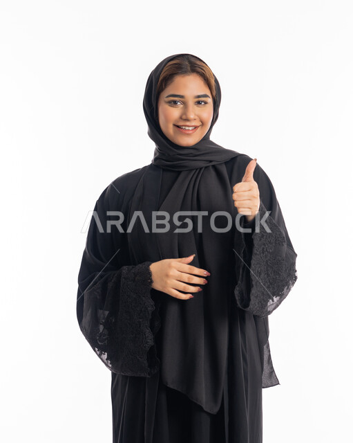 الشعور بحالة جيدة, مفهوم الموافقه والقبول, بورتريه مقرب لامراة عربية خليجية اماراتية مبتسمة محجبة ترتدي العباءة السوداء تشير باصبع الابهام نحو الاعلى بايماءات الجودة و الاعجاب بشيء ما, الاناقة و الاهتمام بالمظهر الخارجي، خلفية بيضاء