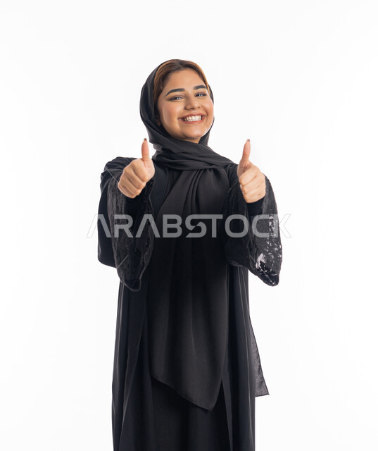 مفهوم الموافقه والقبول, الشعور بحالة جيدة, بورتريه مقرب لامراة عربية خليجية اماراتية مبتسمة محجبة ترتدي العباءة السوداء تشير باصبع الابهام نحو الاعلى بايماءات الجودة و الاعجاب بشيء ما, الاناقة و الاهتمام بالمظهر الخارجي، خلفية بيضاء