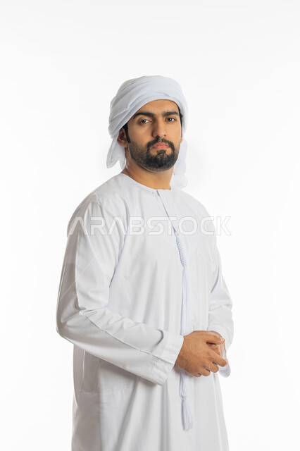 مفهوم الرجولة والقوة والثقة بالنفس، بورتريه من الجانب لرجل عربي خليجي إماراتي يرتدي الكندورة الاماراتية والعمامة ينظر الى الكاميرا بإيماءات الجدية والحزم يقف بأيدي متقاطعة، الاهتمام بالأناقة والمظهر الخارجي، خلفية بيضاء