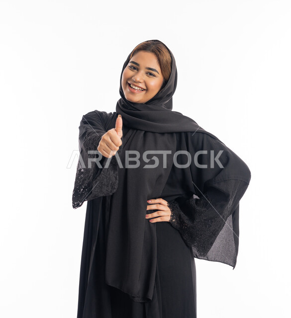 شابة سعودية انيقة، إيماءات الامتياز والجودة، مفهوم القبول والموافقه، بورتريه لامرأه عربيه خليجيه اماراتية محجبة مبتسمة ترتدي عباءة سوداء ترفع اصبع الابهام بتعابير الإعجاب والرضى، خلفية بيضاء