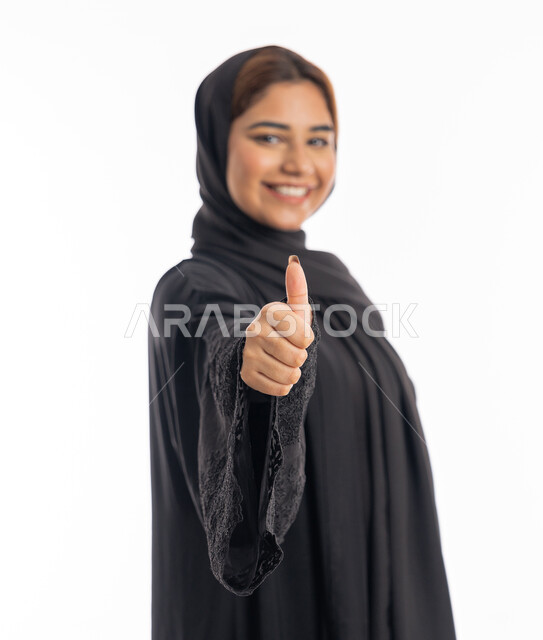 The concept of acceptance and approval, an elegant young Saudi woman, expressions of admiration and satisfaction, a close-up portrait from the side of a smiling veiled Arab Gulf Emirati woman wearing a black abaya, raising her thumb with gestures of excellence and quality, white background.