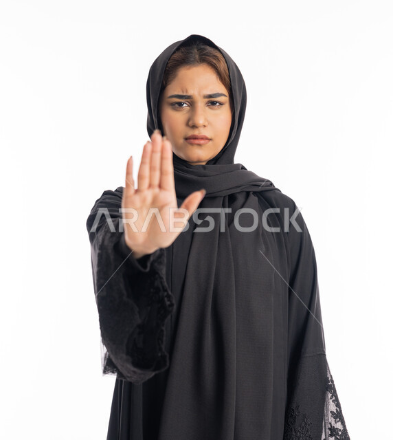 الشعور بالإنزعاج والغضب، شابة سعودية انيقة، تعابير الرفض والإمتناع، الأمر بالتوقف عن فعل شيء ما، بورتريه مقرب لامرأه عربيه خليجيه اماراتية محجة ترتدي عباءة سوداء ترفع يدها بإيماءات تدل على النهي، خلفية بيضاء