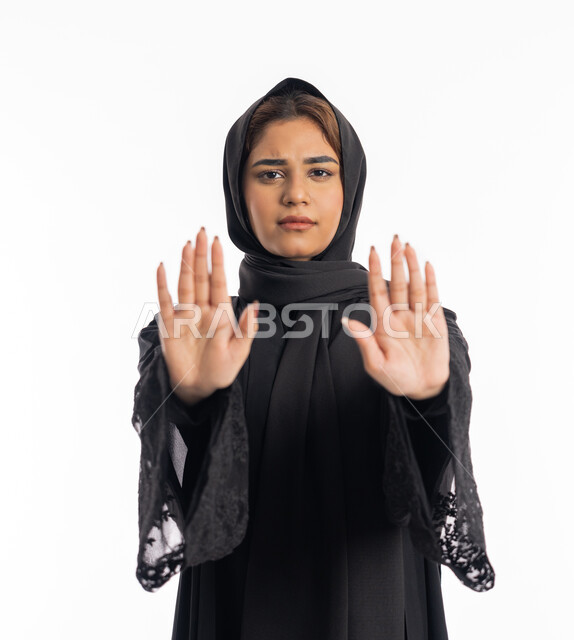 Expressions of rejection and abstention, the command to stop doing something, a close-up portrait of an Arab, Gulf, Emirati, Hajjah woman wearing a black abaya, raising her hands with gestures indicating prohibition, feeling annoyed and angry, an elegant young Saudi woman, white background.