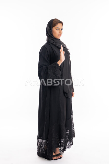 الأمر بالتوقف عن فعل شيء ما، تعابير الرفض والإمتناع، بورتريه من الجانب لامرأه عربيه خليجيه اماراتية محجة ترتدي عباءة سوداء ترفع يدها بإيماءات تدل على النهي، الشعور بالإنزعاج والغضب، شابة سعودية انيقة، صورة لكامل الجسم، خلفية بيضاء
