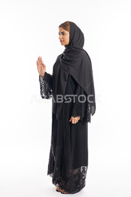Expressions of rejection and abstention, the command to stop doing something, a side portrait of an Arab, Gulf, Emirati, Hajjah woman wearing a black abaya, raising her hand with gestures indicating prohibition, feeling annoyed and angry, an elegant young Saudi woman, full body portrait, white background.