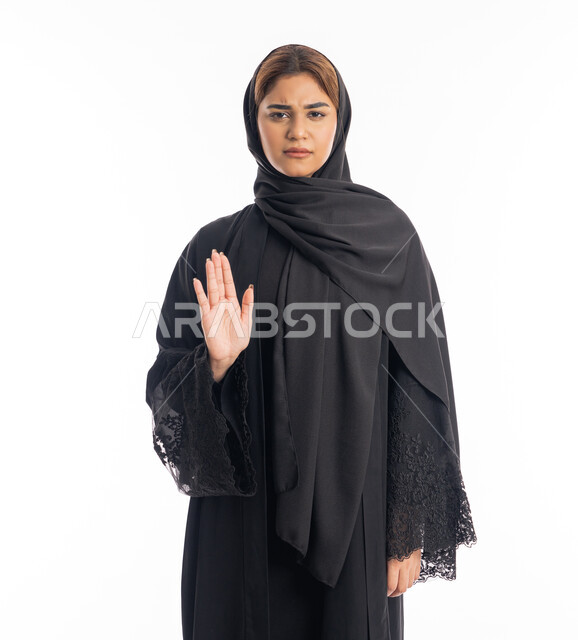 Expressions of rejection and abstention, the command to stop doing something, a portrait of an Arab Gulf Emirati woman from Hajj wearing a black abaya, raising her hands with gestures indicating prohibition, feeling annoyed and angry, an elegant young Saudi woman, white background.