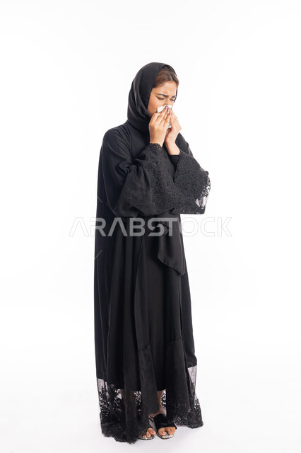 الإصابة بفايروس كورونا كوفيد19، الشعور بالتعب والمرض، استعمال المناديل الورقيه أثناء العطس، بورتريه من الجانب لامرأه عربيه خليجيه اماراتية محجبة ترتدي عباءة سوداء، شابة سعودية انيقة، إيماءات وجه ويدين تدل على العطاس، صورة لكامل الجسم، خلفية بيضاء