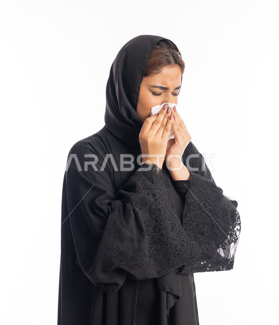 شابة سعودية انيقة، استعمال المناديل الورقيه أثناء العطس، الإصابة بفايروس كورونا كوفيد19، الشعور بالتعب والمرض، بورتريه من الجانب لامرأه عربيه خليجيه اماراتية محجبة ترتدي عباءة سوداء، إيماءات وجه ويدين تدل على العطاس، خلفية بيضاء