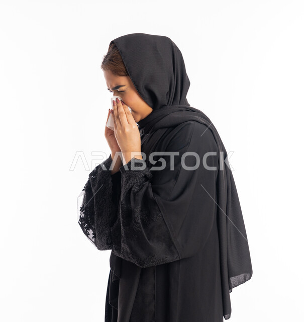 استعمال المناديل الورقيه أثناء العطس، الإصابة بفايروس كورونا كوفيد19، الشعور بالتعب والمرض، بورتريه مقرب من الجانب لامرأه عربيه خليجيه اماراتية محجبة ترتدي عباءة سوداء، شابة سعودية انيقة، إيماءات وجه ويدين تدل على العطاس، خلفية بيضاء