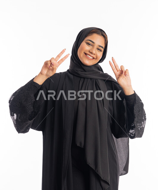 النجاح وتحقيق الأهداف، تعابير السرور والسعادة، بورتريه لامرأة عربية خليجية اماراتية محجبة مبتسمة ترتدي عباءة سوداء تنظر إلى الكاميرا بايماءات وجه ويدين تدل على الفوز والإنتصار، شابة سعودية انيقة، خلفية بيضاء