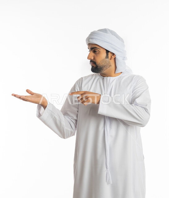حركة أصابع وإشارات، إيماءات حمل ورفع شيء ما، بورتريه لرجل عربي خليجي إماراتي يرتدي الكندورة الإماراتية والعمامة البيضاء يشير باصبع السبابة ليده، الإشارة نحو هدف ما، خلفية بيضاء