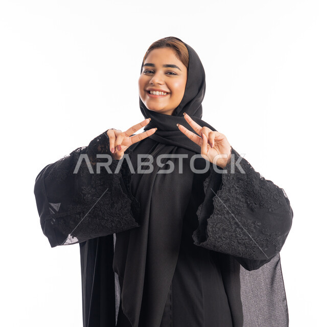 النجاح وتحقيق الأهداف، تعابير السرور والسعادة، بورتريه مقرب لامرأة عربية خليجية اماراتية محجبة مبتسمة ترتدي عباءة سوداء تنظر إلى الكاميرا بايماءات وجه ويدين تدل على الفوز والإنتصار، شابة سعودية انيقة، خلفية بيضاء