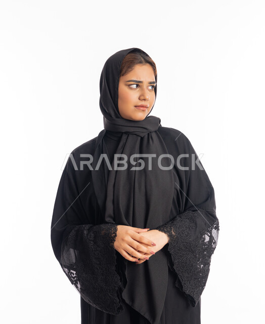 مفهوم الانوثة والاناقة، الاهتمام بالمظهر الخارجي، شابة سعودية تنظر إلى مكان ما بايماءات الحزم والجدية، تعابير الثقة بالنفس، بورتريه مقرب لامرأة عربية خليجية اماراتية محجبة ترتدي عباءة سوداء تقف بأيدي متشبكة، خلفية بيضاء