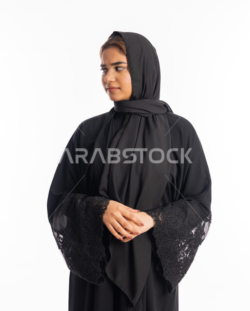 Concern with external appearance, the concept of femininity and elegance, a young Saudi woman looking at a place with gestures of firmness and seriousness, expressions of self-confidence, a close-up portrait of a veiled Arab Gulf Emirati woman wearing a black abaya, standing with clasped hands, white background.