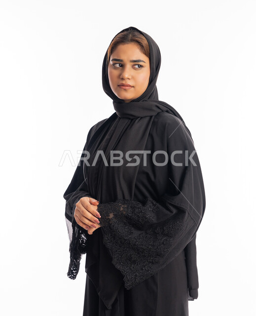 مفهوم الانوثة والاناقة، الاهتمام بالمظهر الخارجي، شابة سعودية تنظر إلى مكان ما بايماءات الحزم والجدية، تعابير الثقة بالنفس، بورتريه من الجانب لامرأة عربية خليجية اماراتية محجبة ترتدي عباءة سوداء تقف بأيدي متشبكة، خلفية بيضاء