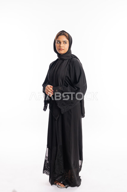الاهتمام بالمظهر الخارجي، شابة سعودية تنظر إلى مكان ما بايماءات الحزم والجدية، تعابير الثقة بالنفس، بورتريه من الجانب لامرأة عربية خليجية اماراتية محجبة ترتدي عباءة سوداء تقف بأيدي متشبكة، صورة الجسم بالطول الكامل، خلفية بيضاء