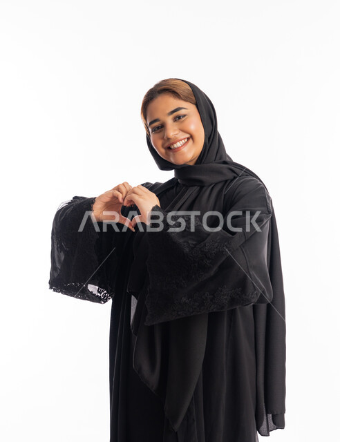 تعابير السرور والسعادة، ايماءات يدين على شكل قلب، اظهار مشاعر الحب والشغف، مفهوم المحبة والمودة، بورتريه من الجانب لامرأه عربيه خليجيه اماراتية محجبة ترتدي عباءة سوداء تنظر إلى الكاميرا بتعابير السرور والسعادة، شابة سعودية انيقة، خلفية بيضاء