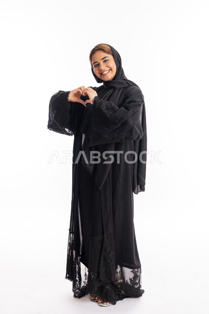 Heart-shaped hand gestures, showing feelings of love and passion, the concept of love and affection, a portrait from the side of a veiled Arab Gulf Emirati woman wearing a black abaya looking at the camera with expressions of pleasure and happiness, an elegant young Saudi woman, full body portrait, white background