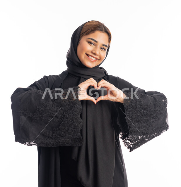 ايماءات يدين على شكل قلب، اظهار مشاعر الحب والشغف، مفهوم المحبة والمودة، بورتريه مقرب لامرأه عربيه خليجيه اماراتية محجبة ترتدي عباءة سوداء تنظر إلى الكاميرا بتعابير السرور والسعادة، شابة سعودية انيقة، خلفية بيضاء