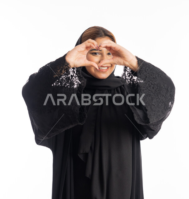 اظهار مشاعر الحب والشغف، مفهوم المحبة والمودة، ايماءات يدين على شكل قلب، بورتريه مقرب لامرأه عربيه خليجيه اماراتية محجبة ترتدي عباءة سوداء تنظر إلى الكاميرا بتعابير السرور والسعادة، شابة سعودية انيقة، خلفية بيضاء