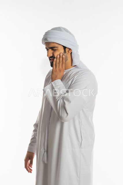 تسوس الأضراس وإلتهاب اللثه، طلب العلاج والمساعده، بورتريه لرجل عربي خليجي اماراتي يرتدي الكندورة الاماراتية والعمامة البيضاء يضع يده على خده بإيماءات تدل على الشعور بألم الأسنان، الحاجه لزيارة الطبيب، خلفية بيضاء