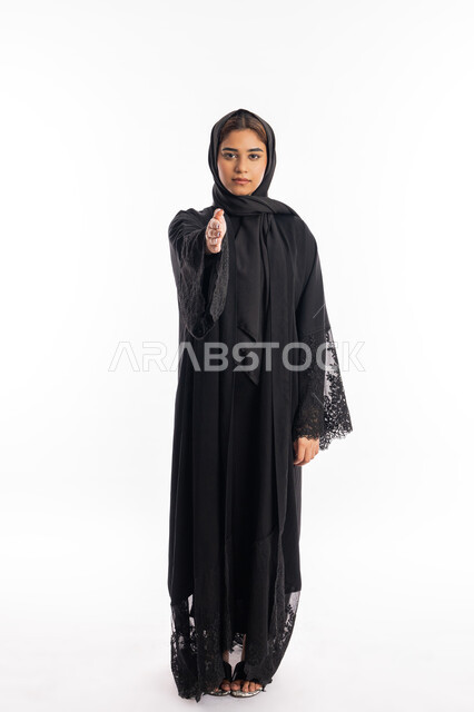 إلقاء التحيه والسلام، مفهوم الاستقبال والترحيب، شابة سعودية تنظر إلى الكاميرا بإيماءات الجدية، التعارف وتكوين الصداقات، بورتريه لامرأة عربية خليجية إماراتية ترتدي العباءة السوداء تقوم بمد كفها للمصافحة، صورة لكامل الجسم، خلفية بيضاء