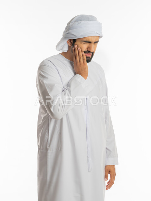 الحاجه لزيارة الطبيب، تسوس الأضراس وإلتهاب اللثه، طلب العلاج والمساعده، بورتريه لرجل عربي خليجي اماراتي يرتدي الكندورة الاماراتية والعمامة البيضاء يضع يده على خده بإيماءات تدل على الشعور بألم الأسنان، خلفية بيضاء