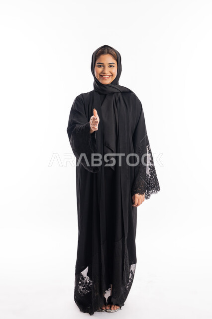 Standing confidently and looking at the camera, satisfaction and pleasure in getting to know each other and forming successful relationships, the concept of greetings and peace, portrait of a smiling Arab Gulf Emirati young woman wearing the black abaya, standing with positivity and happiness, a Saudi woman extending her palm to shake hands, full body portrait, white background