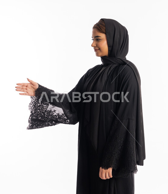مفهوم الاستقبال والترحيب, إلقاء التحيه والسلام, سيدة سعودية تقوم بالتعارف و الصداقه وتكوين علاقات, بورتريه من الجانب لشابة عربية خليجية إماراتية ترتدي العباءة السوداء تقوم بمد كفها للمصافحة، خلفية بيضاء