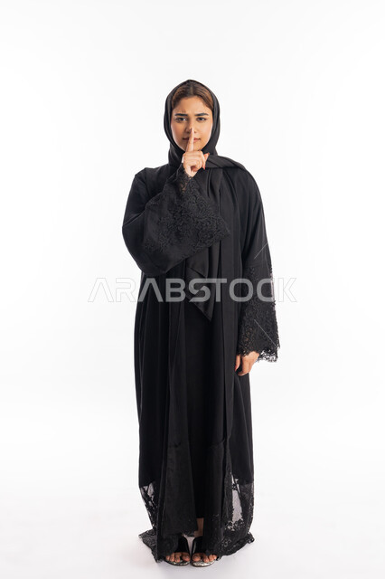 سيدة سعودية تضع أصبع السبابة على فمها بإيماءات عدم التدخل وطلب السكوت والصمت, بورتريه لشابة عربية خليجية إماراتية ترتدي العباءة السوداء تقف بجدية وتنظر إلى الكاميرا, الهدوء والسكون وعدم الإزعاج، التوقف عن الكلام وخفض الصوت, صورة الجسم بالكامل, خلفية بيضاء