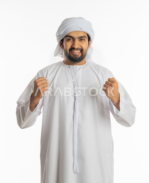 تحقيق الاهداف و إنجاز المهام, الوقوف بسرور والنظر إلى الكاميرا, علامات وتعابير والفرح والقوة, الإعجاب والرضا و الشعور بالسعاده، بورتريه لرجل عربي خليجي اماراتي مبتسم يرتدي العمامة والكندورة الاماراتية يرفع يديه بإيماءات النصر والفوز و الكسب, خلفية بيضاء