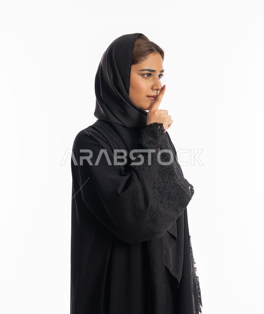 الهدوء والسكون وعدم الإزعاج، التوقف عن الكلام وخفض الصوت, بورتريه من الجانب لشابة عربية خليجية إماراتية ترتدي العباءة السوداء تقف بجدية, سيدة سعودية تضع أصبع السبابة على فمها بإيماءات عدم التدخل وطلب السكوت والصمت, خلفية بيضاء
