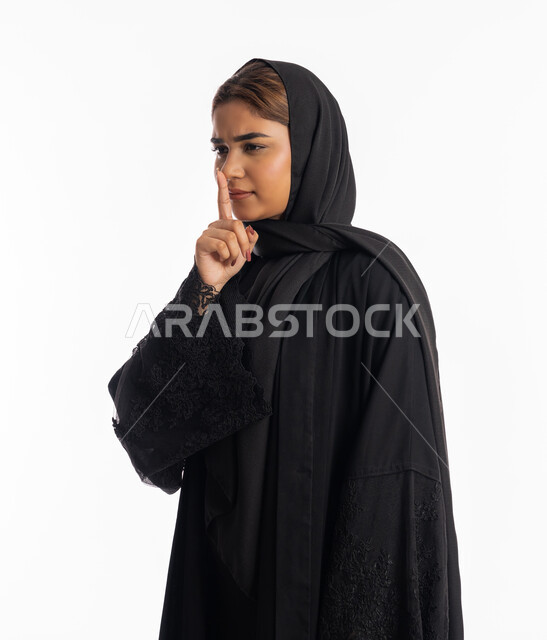 التوقف عن الكلام وخفض الصوت, الهدوء والسكون وعدم الإزعاج، بورتريه من الجانب لشابة عربية خليجية إماراتية ترتدي العباءة السوداء تقف بجدية, سيدة سعودية تضع أصبع السبابة على فمها بإيماءات عدم التدخل وطلب السكوت والصمت, خلفية بيضاء