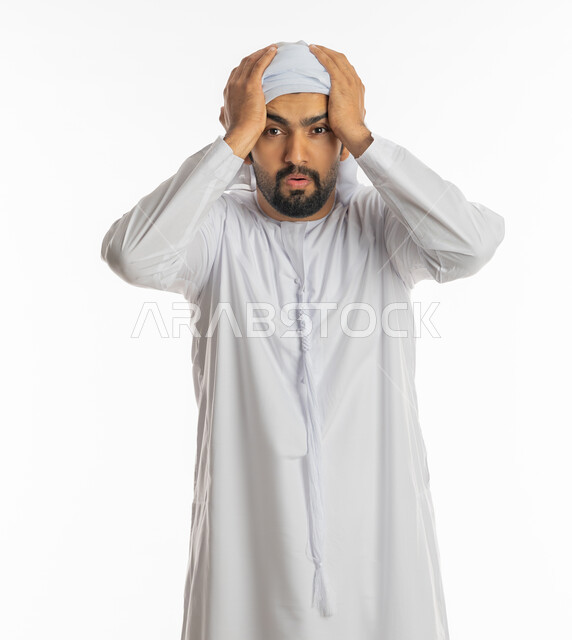 Astonishment and disbelief, loss and loss of something specific, surprise and astonishment at something, standing with gestures of astonishment and astonishment, portrait of a young Arab Gulf Emirati man wearing a kandora and an Emirati turban, raising his hands on his head with an expression of shock and astonishment, white background.
