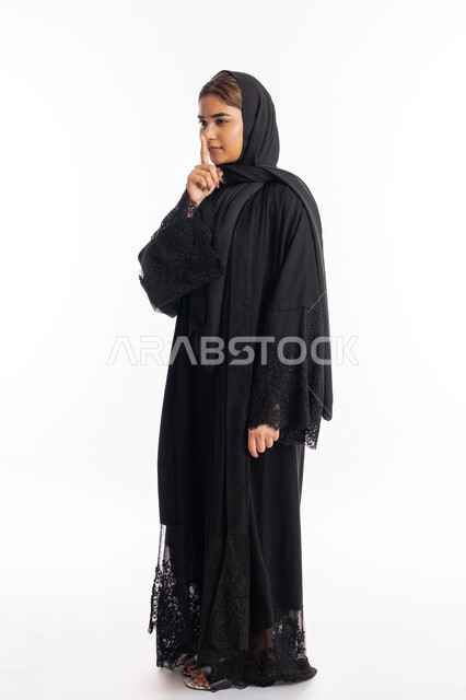 الوقوف بجدية والأمر بالتوقف عن الكلام وخفض الصوت, الهدوء والسكون وعدم الإزعاج، بورتريه لإمرأة عربية خليجية إماراتية ترتدي العباءة السوداء تضع أصبع السبابة على فمها, سيدة سعودية تقف بإيماءات عدم التدخل وطلب السكوت والصمت, صورة الجسم بالكامل, خلفية بيضاء