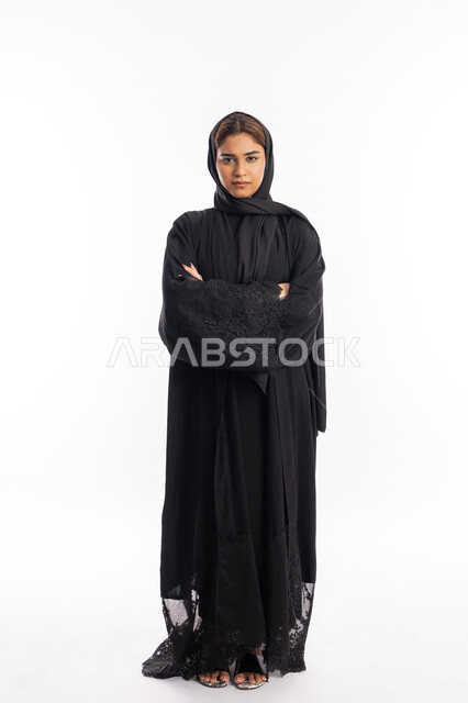 الاهتمام والعنايه بالمظهر الخارجي، شابة سعودية تقف بجدية وبأيدي متقاطعة، مفهوم الأنوثة والنعومة، بورتريه لامرأة عربية خليجية اماراتية محجبة ترتدي العباءة السوداء تنظر إلى الكاميرا بايماءات الثقة بالنفس، صورة لكامل الجسم، خلفية بيضاء