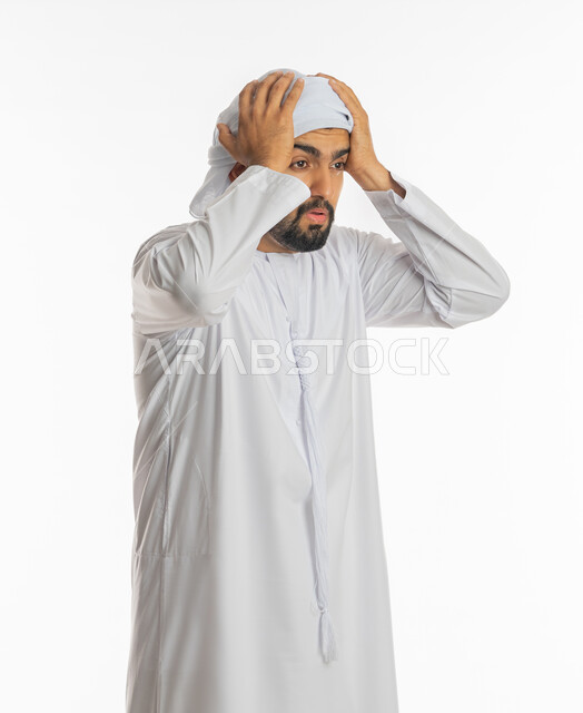 Standing with gestures of astonishment and astonishment, surprise and astonishment at something, portrait of a young Arab Gulf Emirati man wearing a kandura and an Emirati turban, raising his hands on his head with expressions of shock and astonishment, astonishment and disbelief at a certain matter, white background.